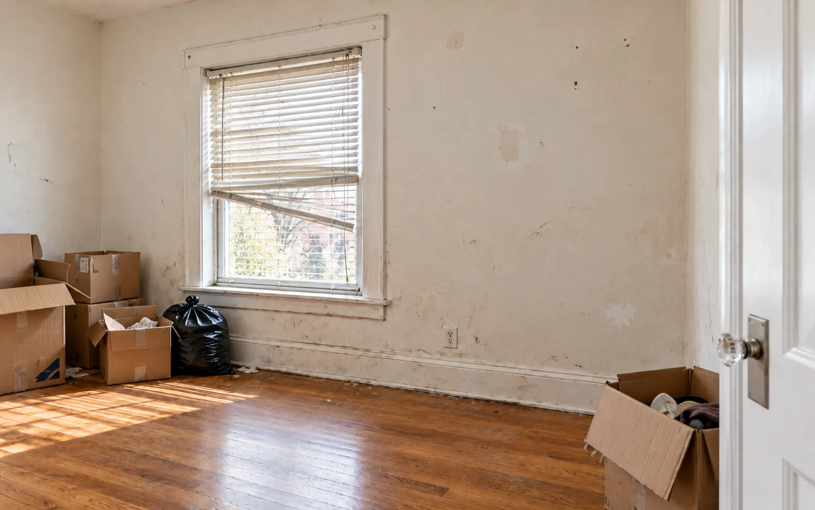 A room in move-out condition with scuffed walls, a broken blind and boxes
